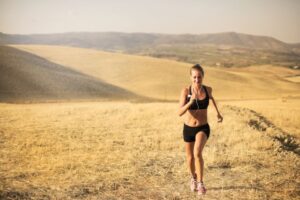 Powerful female athlete in sportswear and earphones jogging through field in sunny hot day